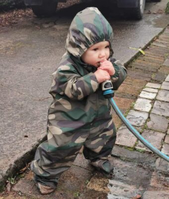 Toddler playing outside with a hose, keeping clean and dry as he's wearing Mud Mates NZ-made waterproof overalls and shoe covers. Save money with a Busy Crawler Pack
