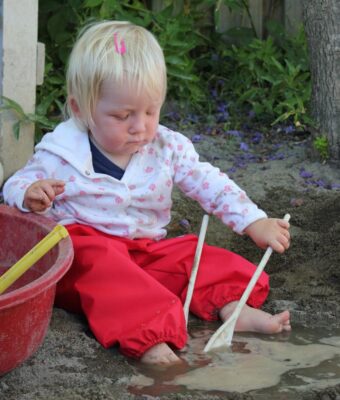 Baby wearing red overpants, sitting in the sandpit with water