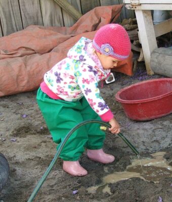 Young child wearing green overpants playing with a hose in the sandpit