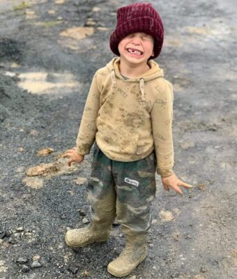 Child with a big grin on his face and very muddy Overpants in green camo