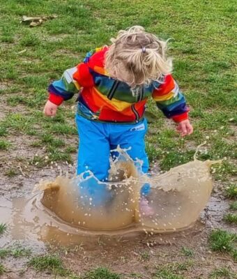Child in turquoise overpants splashing in a muddy puddle