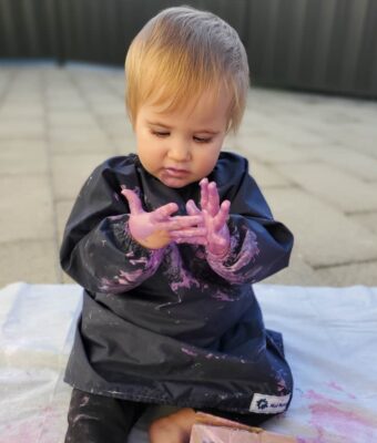 Toddler wearing a Mud Mates NZ-made navy blue craft apron/art smock with paint on her hands