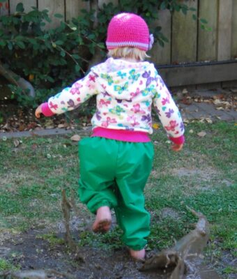 Young girl splashing in a muddy puddle, keeping clean and dry wearing Mud Mates NZ-made waterproof overpants in green