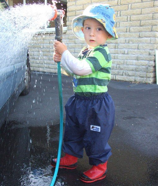 Young boy helping wash a car, playing with hose and water, keeping dry wearing Mud Mates NZ-made waterproof overpants in navy