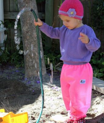 Young girl in sandpit playing with hose and water wearing Mud Mates NZ-made waterproof overpants in pink