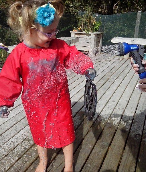 Young child wearing a paint covered Mud Mates Craft Apron being sprayed with water