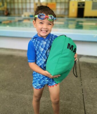 Young boy in his togs holding a green Mud Mates swim bag