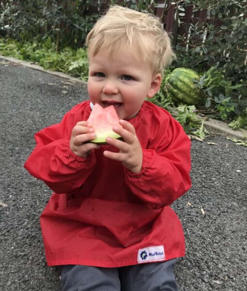 Toddler sitting and eating a slice of watermelon, his clothes protected by a red Mud Mates Long Sleeve Bib