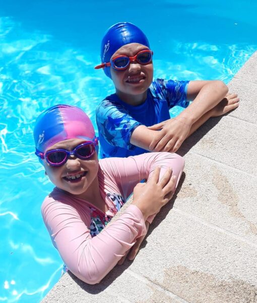 A girl and boy at edge of swimming pool wearing silicone kids swim caps and Funkies kids goggles