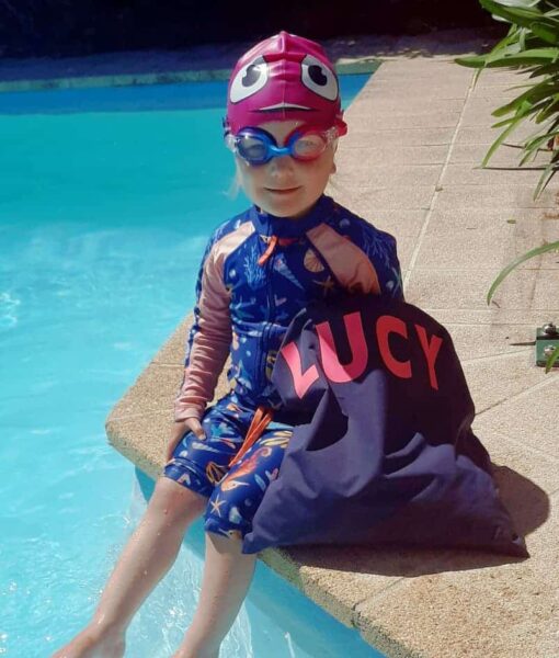 Toddler girl sitting by swimming pool with her personalised NZ-made swim bag, purple/pink kids goggles and pink fish swim cap