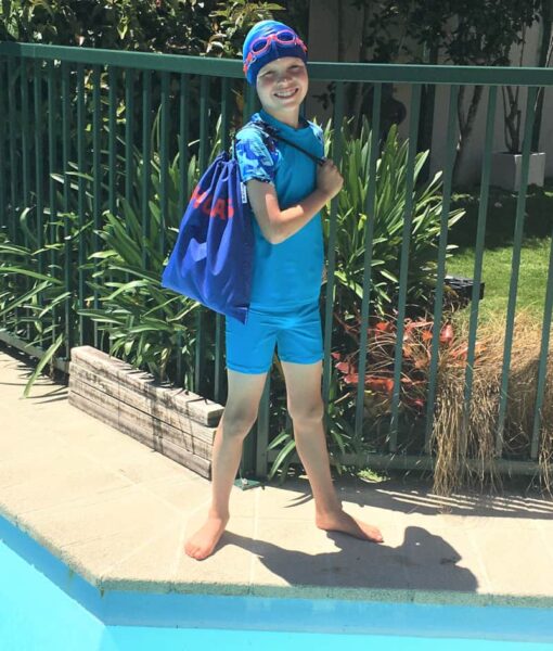 Boy standing by pool wearing swim cap and goggles and holding a Mud Mates NZ-made personalised swim bag