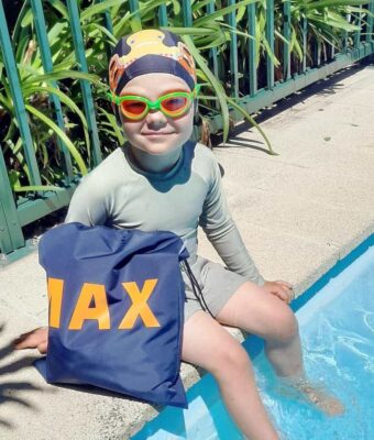 Young boy sitting by swimming pool with his personalised NZ-made swim bag, orange and green goggles and lycra octopus swim cap