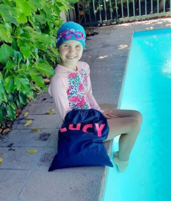 Young girl sitting by swimming pool with her personalised NZ-made swim bag, pink/purple kids goggles and daisy chain lycra swim cap