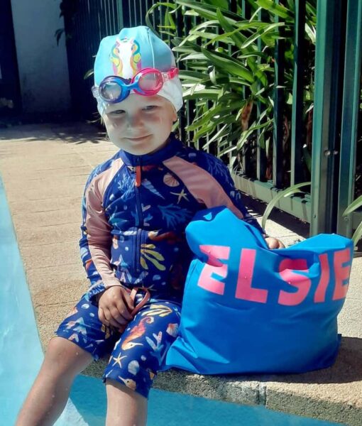 Toddler girl sitting by swimming pool with her personalised NZ-made swim bag, kids goggles and swim cap