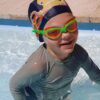 Young happy boy in swimming pool wearing Otto the Octopus lycra kids swim cap and orange/green Funkies kids goggles