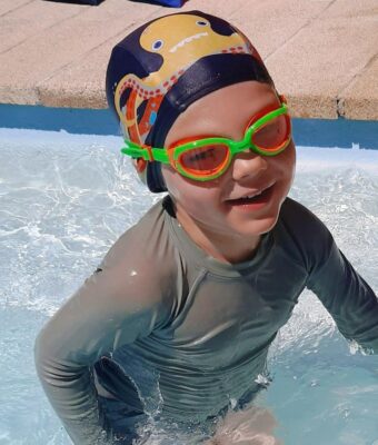Young happy boy in swimming pool wearing Otto the Octopus lycra kids swim cap and orange/green Funkies kids goggles