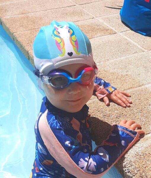 Toddler in swimming pool wearing rainbow drop unicorn lycra kids swim cap and goggles