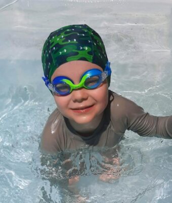Toddler in swimming pool wearing a lycra swim cap and rainbow blue/green kids goggles