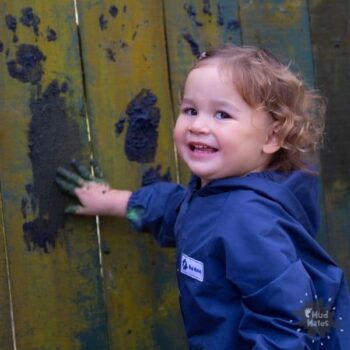 Young girl wearing Mud Mates NZ-made coveralls mud painting with her fingers on a fence