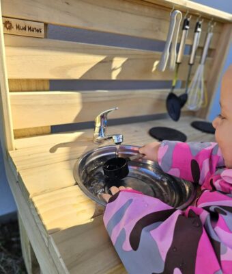 Mud Mates NZ-made wooden mud kitchens feature actual running water! Young girl plays with the water, keeping dry in her waterproof Coveralls