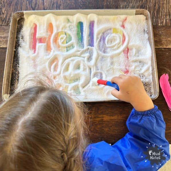 Young child using a paintbrush to write 'hello' in the salt using a rainbow salt writing tray