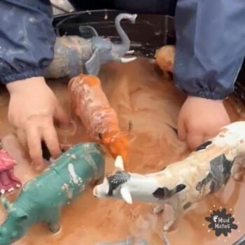 Child's hands playing in a sensory bin with muddy oobleck / cornflour gloop and toy animals.