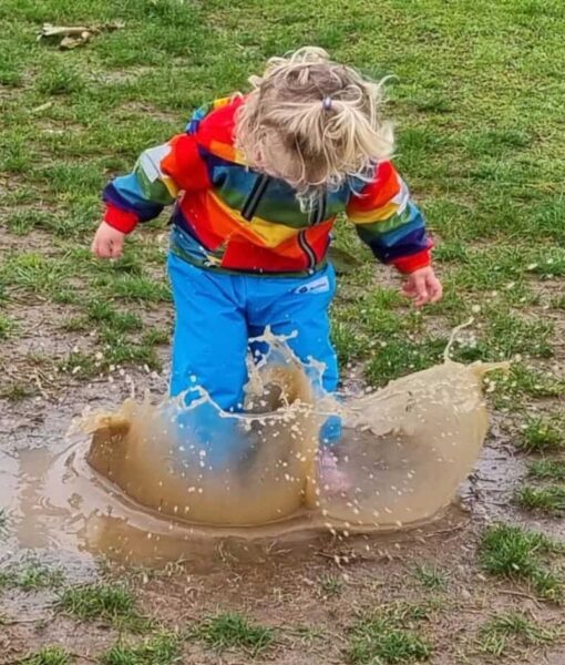 Young child jumping in a muddy puddle, keeping clean and dry in her Mud Mates waterproof overpants