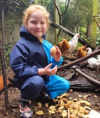 Young girl sitting with chickens holding an egg, wearing navy/turquoise children's coveralls