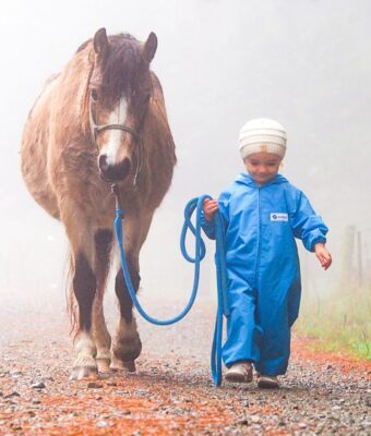 Young child walking a horse in the fog, keeping clean and dry in her turquoise waterproof children's coveralls