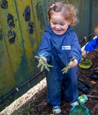 Young girl enjoying mud painting - laughing at her muddy hands - keeping clean and dry in her Mud Mates children's coveralls