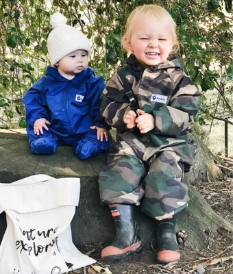 Two young children wearing waterproof coveralls, one in royal blue and one in camo, sitting outdoors