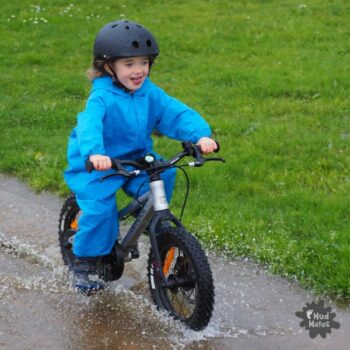 Fun & Free Kids Activities for the School Holidays - young girl riding her bike through a puddle of water, keeping dry in her Mud Mates coveralls