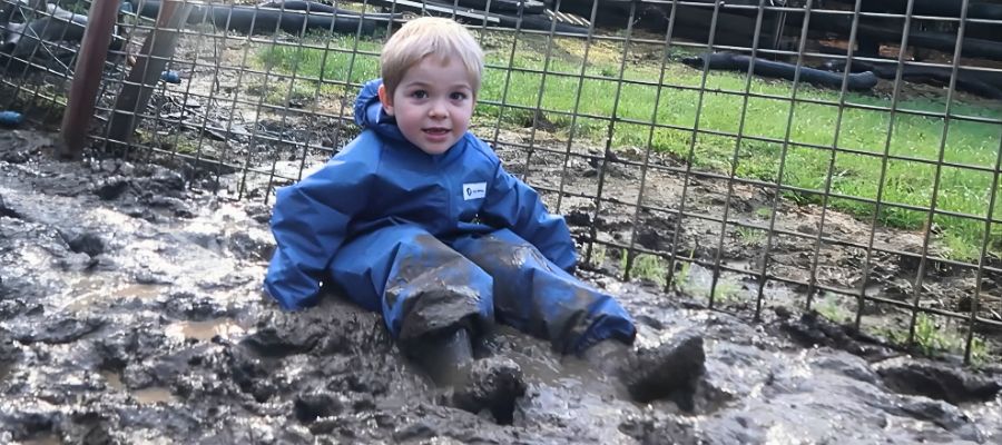 Boy sitting in a pile of mud in blue coveralls enjoying the benefits of muddy play