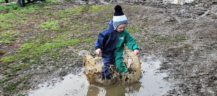 Boy jumping in a muddy puddle wearing coveralls, illustrating the benefits of muddy play