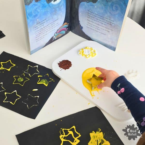 Girl stamping Matariki stars onto dark card with cookie cutters and paint alongside a Matariki book